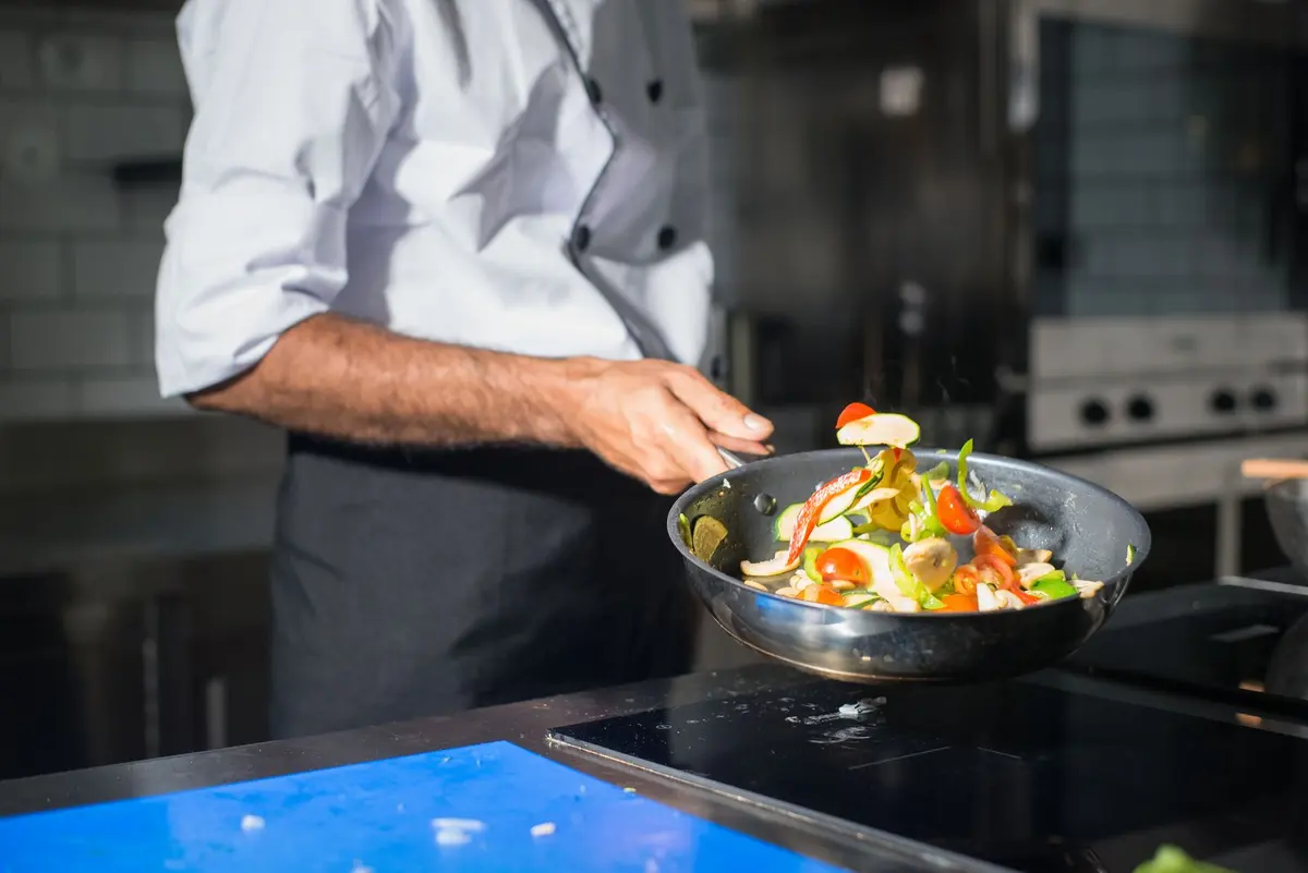 Hands tossing fresh vegetables in a frying pan over a stove in a bright kitchen.
